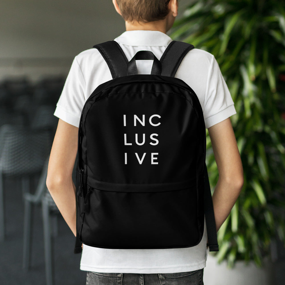 This is a photo of URevolution's black Inclusive backpack. In the top half of the backpack, the word inclusive is printed in white upper case letters evenly spaced over three lines: INC LUS IVE. The rest of the backpack is black. The backpack is work by a teenager wearing a white polo shirt and black jeans.
