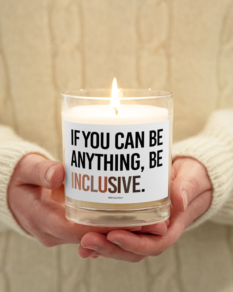 A Be Inclusive Diversity candle. On the front of the diversity candle is the phrase: "If you can be anything, be inclusive," in upper case letters printed on a white label. The text is all black, except the word "Inclusive," which is in different skin colors, with the lightest color on the left and the darkest on the right. The candle is being held in a person's hands.