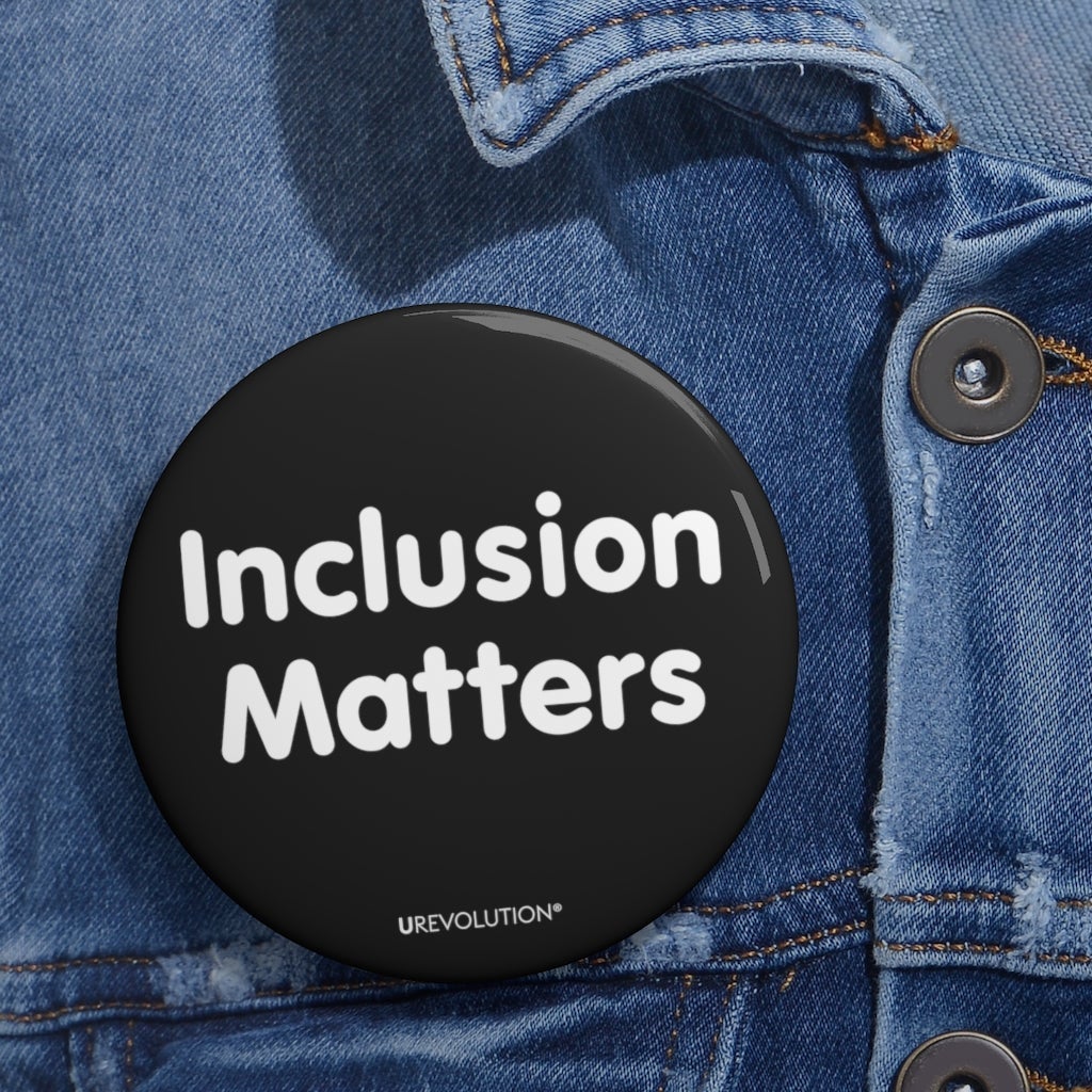 This is a photo of a 3" diameter black Inclusion Matters pin on a blue denim jacket. A black Inclusion Matters pin button against a plain white background. On the front of the pin button are two words, 'Inclusion Matters.' printed in white sentence case letters over two lines. At the bottom edge of the inclusion badge, beneath the phrase, 'Inclusion Matters,' is the brand name URevolution in small white upper case letters.