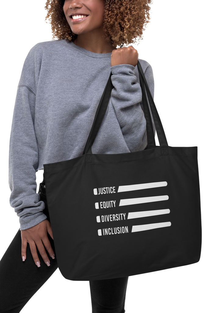 This is a large black Justice Equity Diversity Inclusion organic tote bag. Four white laser swords are placed horizontally in the middle of the tote, similar to a flag. On the left-hand side of each sword, in the place of the handle, is one word: Justice Equity Diversity Inclusion. The tote is being held by a model.
