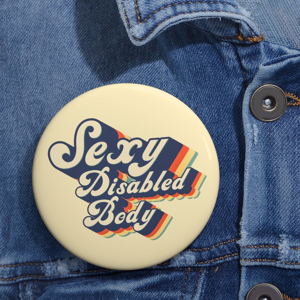 This is a photo of a 3" URevolution's round Sexy Disabled Body pin badge on a blue denim jacket. The pin is a light cream color and features the phrase, Sexy Disabled Body, in a folkloric font.