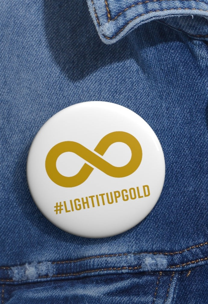 This is a photo of a Light It Up Gold Pin Button. The pin has the word #LightItUpGold printed in large upper case letters beneath a solid line infinity symbol. The symbol and text are colored gold, with the background being soft-white. The pin is pinned on a faed blue denim jacket.