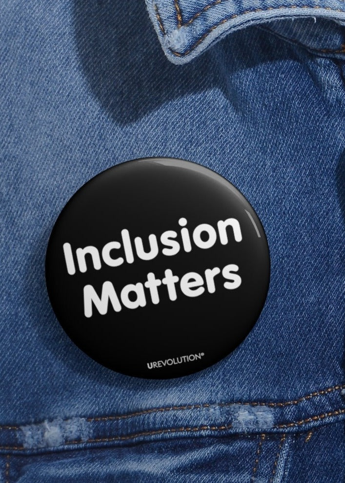This is a photo of a 2.25" diameter black Inclusion Matters pin on a blue denim jacket. A black Inclusion Matters pin button against a plain white background. On the front of the pin button are two words, 'Inclusion Matters.' printed in white sentence case letters over two lines. At the bottom edge of the inclusion badge, beneath the phrase, 'Inclusion Matters,' is the brand name URevolution in small white upper case letters.