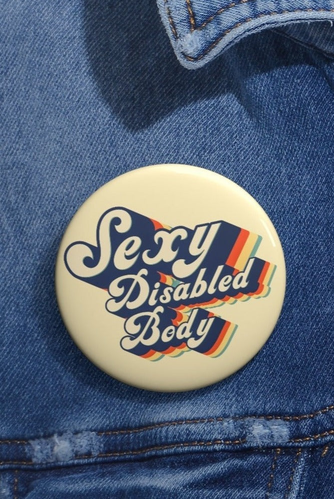 This is a photo of a 2.25" URevolution's round Sexy Disabled Body pin badge on a blue denim jacket. The pin is a light cream color and features the phrase, Sexy Disabled Body, in a folkloric font.