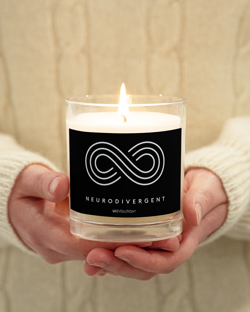 Neurodivergent candle by URevolution. This is a photo of a candle in a glass jar with a black label. In the middle of the black label is a three-ringed infinity symbol with the word 'neurodivergent' in white upper case letters beneath the symbol.  The candle is held between two hands.