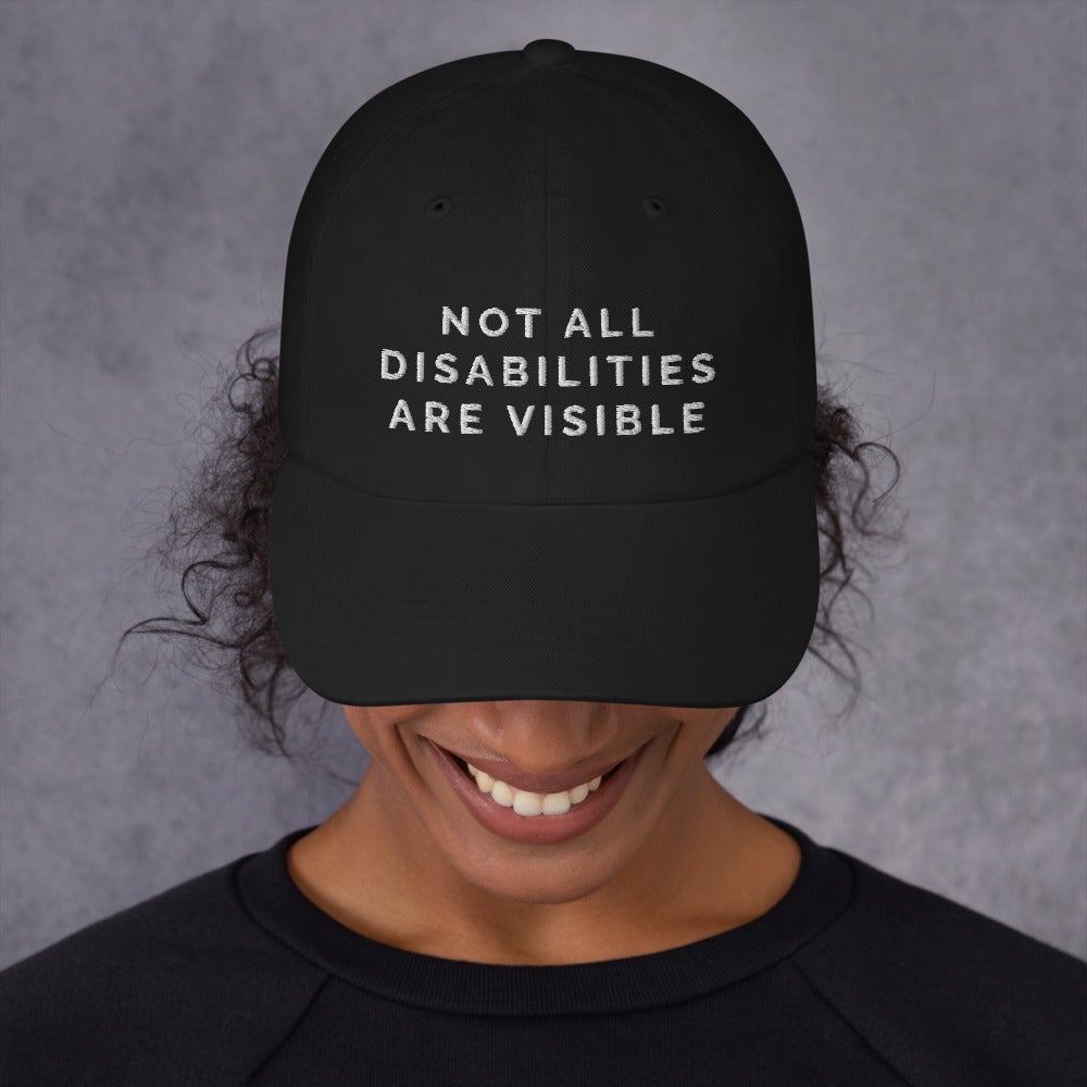This is a photo of the front of a person's face wearing a black Not All Disabilities Are Visible Cap. In the middle of the cap, in black embroidered upper case letters, is the phrase "Not All Disabilities Are Visible." The phrase is on three lines: (1) not all, (2) disabilities, (3) are visible. The person's face is not visible except for a smile and long-hair beneath the cap.