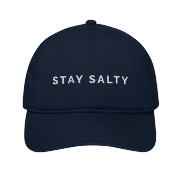 A photo of a navy-pacific colored organic Stay Salty cap lying flat against a plain background. On the front panel of the cap the phrase, 'Stay Salty,' is embroidered on it in white upper case letters. 