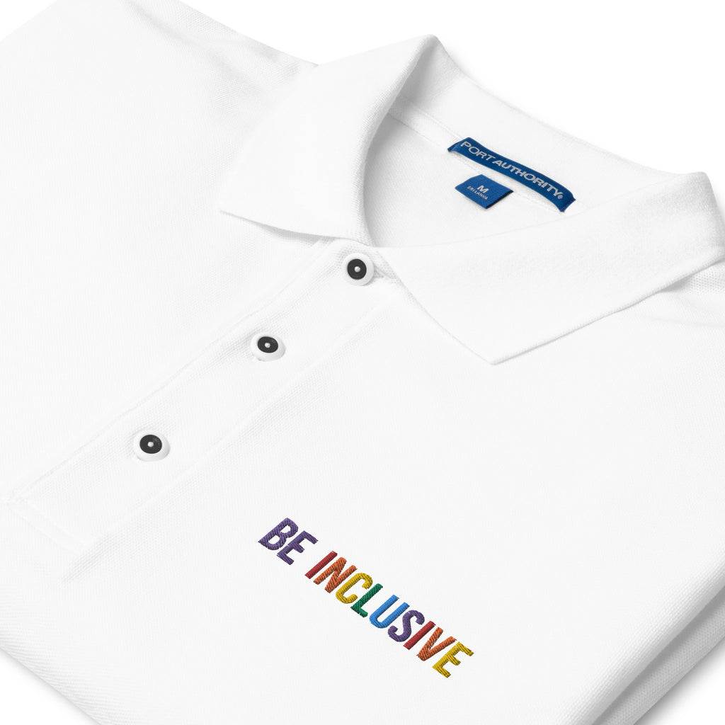This is a photo of a premium white Rainbow Polo Shirt. On the polo's top right panel is the phrase, Be Inclusive, embroidered in red, orange, yellow, green, indigo, and violet. The word 'be' is only in violet. Close up of the polo collar is shown.