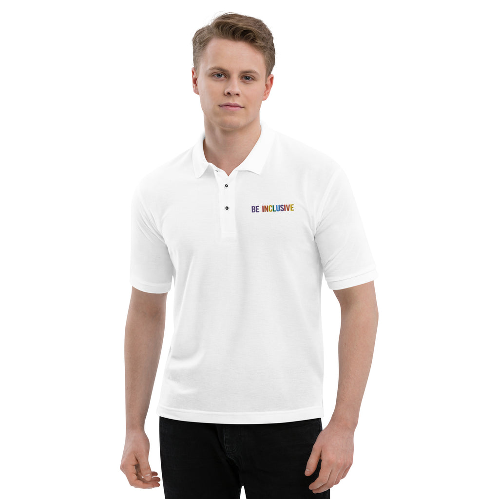 Photo of a premium white Rainbow Be Inclusive Polo Shirt. On the polo's top right panel is the phrase, Be Inclusive, embroidered in red, orange, yellow, green, indigo, and violet. The word 'be' is only in violet. The polo is won by a male model.