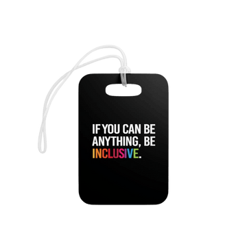 This is a close-up photo of a black rectangle be inclusive luggage tags on a wooden desk. The luggage tag has the phrase 'if you can be anything, be inclusive' in the middle of the tag in white upper case letters. The word inclusive is printed in the colors of the rainbow.