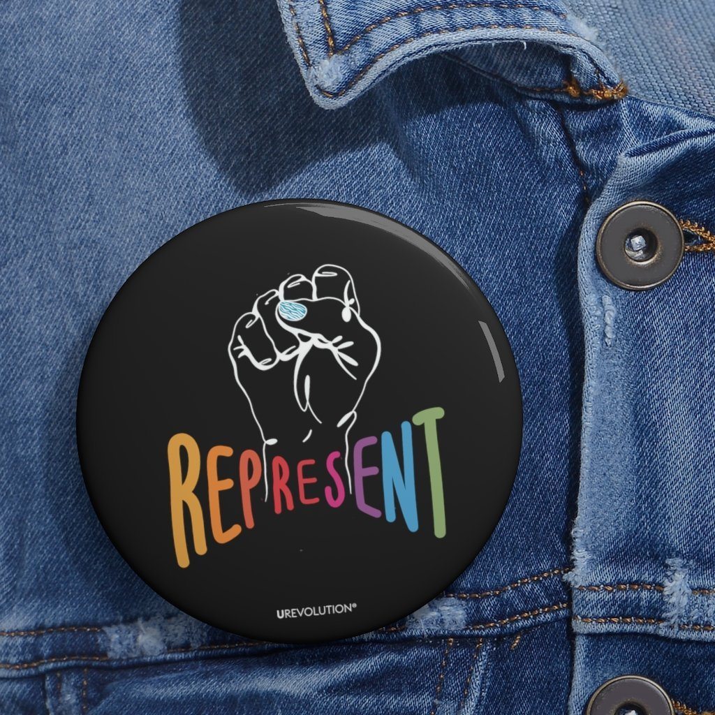 Represent pin button. The round black pin button is pinned on a slightly faded denim jacket. In the middle of the pin is a white drawing of a raised clenched fist, with the handwritten word “represent,” written in upper case rainbow colors.
