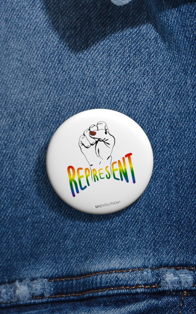 Represent Pride Pin Button. In the middle of the represent pride pinback button is a black line drawing of a raised clenched fist, with the handwritten word "represent," written in blended upper case rainbow pride colors: red, orange, yellow, green, indigo, and violet. Under the fist, at the bottom of the magnet, is the word URevolution in small black upper case letters.. The 1.25 inch pin button is displayed on the front of a faded blue denim jacket.