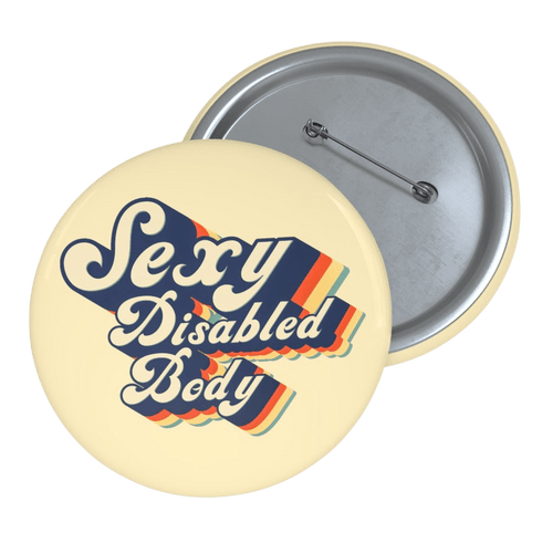 This is a photo of a 1.25" URevolution's round Sexy Disabled Body pin badge on a blue denim jacket. The pin is a light cream color and features the phrase, Sexy Disabled Body, in a folkloric font.
