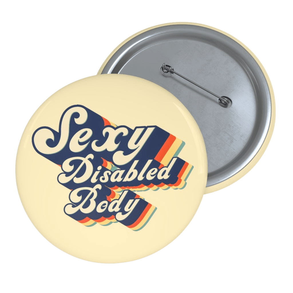 This is a photo of a 1.25" URevolution's round Sexy Disabled Body pin badge on a blue denim jacket. The pin is a light cream color and features the phrase, Sexy Disabled Body, in a folkloric font.