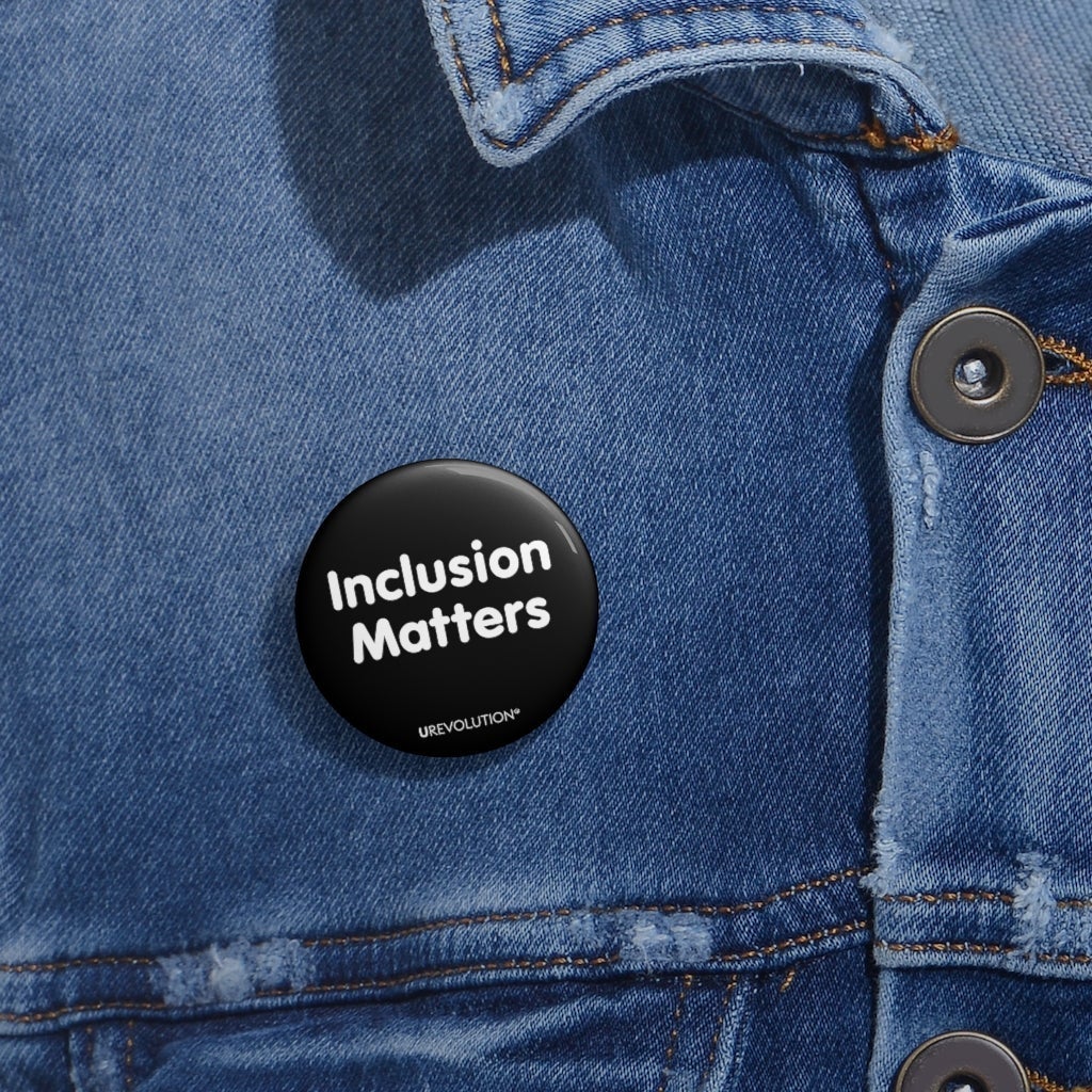 A black Inclusion Matters pin button against a plain white background. On the front of the pin button are two words, 'Inclusion Matters.' printed in white sentence case letters over two lines. At the bottom edge of the inclusion badge, beneath the phrase, 'Inclusion Matters,' is the brand name URevolution in small white upper case letters. The small  Inclusion Matters pin button badge is worn on a blue denim jacket. 