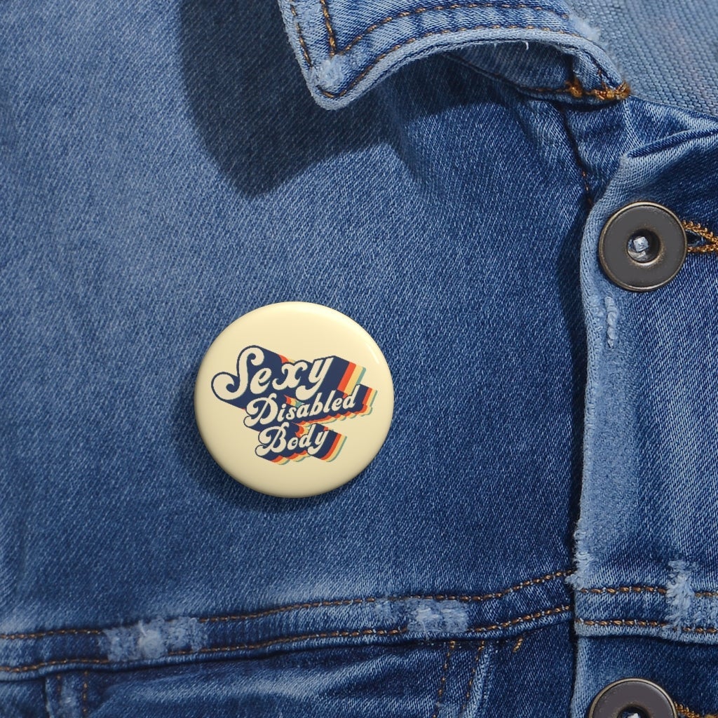 This is a photo of a 1.25" URevolution's round Sexy Disabled Body pin badge on a blue denim jacket. The pin is a light cream color and features the phrase, Sexy Disabled Body, in a folkloric font.