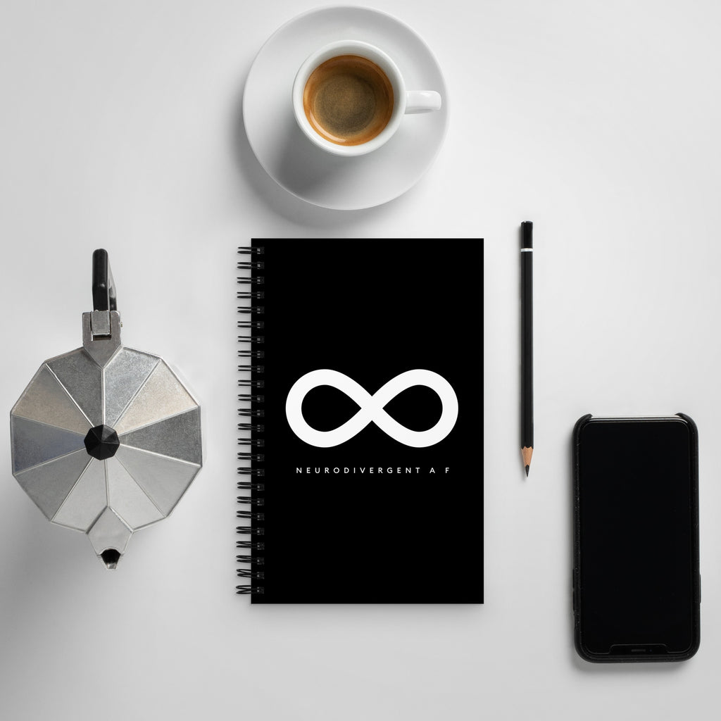 This is a photo of a spiral black Neurodivergent AF notebook. In the middle of the black notebook is an elegant black infinity symbol consisting of three thin black lines. Just below the infinity symbol is the word Neurodivergent AF in elegant upper case white letters. The notebook is on a table.