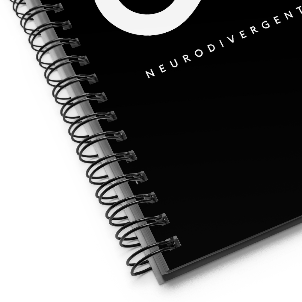 This is a photo of a spiral black Neurodivergent AF notebook. In the middle of the black notebook is an elegant black infinity symbol consisting of three thin black lines. Just below the infinity symbol is the word Neurodivergent AF in elegant upper case white letters. This is a close up of the bottom left-hand corner of the notebook.