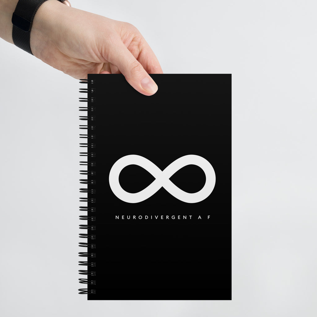This is a photo of person's hand holding a spiral black Neurodivergent AF notebook. In the middle of the black notebook is an elegant black infinity symbol consisting of three thin black lines. Just below the infinity symbol is the word Neurodivergent AF in elegant upper case white letters. [Neurodivergent AF means Neurodivergent As Fuck]