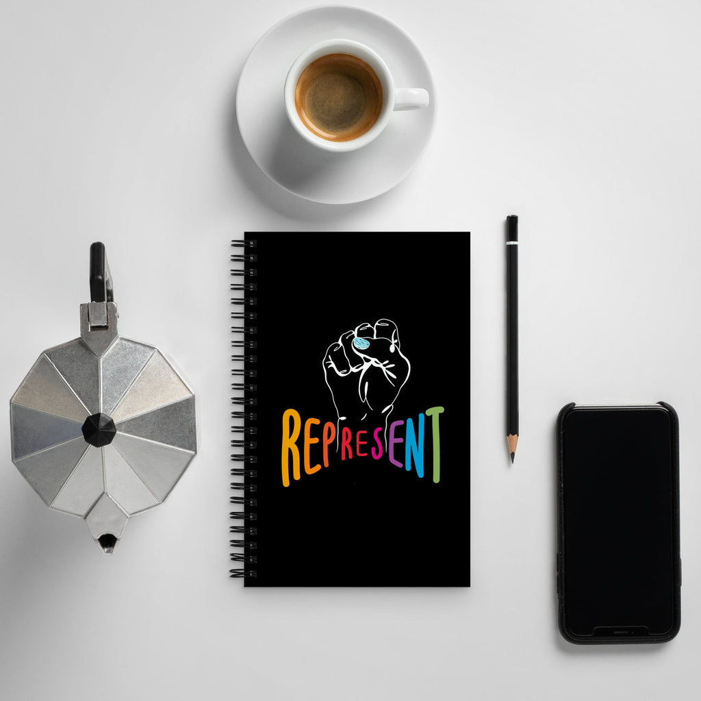 This is a photo of spiral black Represent notebook. In the center of the notebook is a white drawing of a raised clenched fist, with the handwritten word "represent," written in upper case rainbow colors beneath the fist. The represent notebook is on a coffee table.