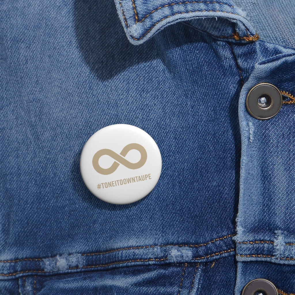  This is a photo of a Tone It Down Taupe Pin. The pin has the word #ToneItDownTaupe printed in large upper case letters beneath a solid line infinity symbol. The symbol and text are colored taupe, with the background being soft-white. The 1.25 inch pin is pinned to a faded blue denim jacket. 