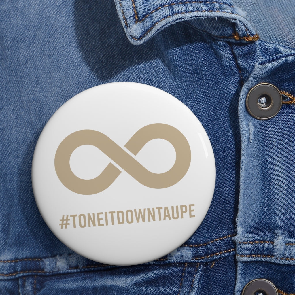  This is a photo of a Tone It Down Taupe Pin. The pin has the word #ToneItDownTaupe printed in large upper case letters beneath a solid line infinity symbol. The symbol and text are colored taupe, with the background being soft-white. The 3 inch pin is pinned to a faded blue denim jacket. 