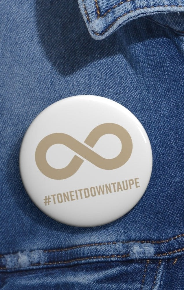  This is a photo of a Tone It Down Taupe Pin. The pin has the word #ToneItDownTaupe printed in large upper case letters beneath a solid line infinity symbol. The symbol and text are colored taupe, with the background being soft-white. The 2.25 inch pin is pinned to a faded blue denim jacket. 