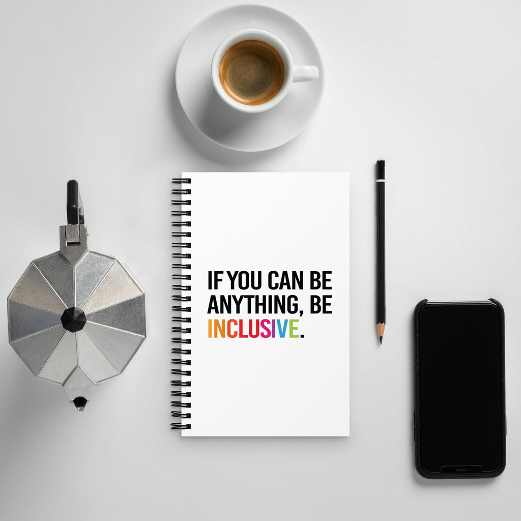 Be Inclusive Journal. The front cover of a white spiral journal with the following phrase printed in the middle of it in upper case letters: 'If you can be anything, be inclusive.' The word inclusive is in the color of the rainbow.  The journal is placed on a table next to a coffee cup and phone.