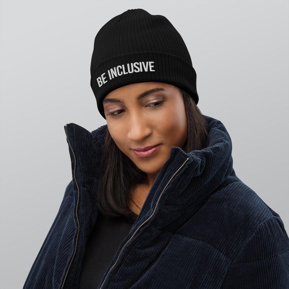 This is a photo of a person standing against a white background wearing a black jacket and a black Be Inclusive organic beanie. The words 'Be Inclusive' are embroidered on the front of the beanie in white upper case letters.