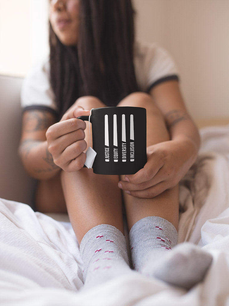 A black J.E.D.I. Justice Equity Diversity Inclusion Mug. In the middle of the mug are four white laser swords. At the bottom of each sword representing the handle is one word: Justice Equity Diversity Inclusion.  A woman is sitting in bed with the mug.